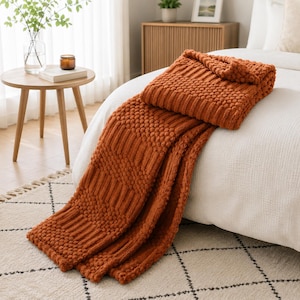 May include: A burnt orange throw blanket with a textured pattern featuring alternating sections of raised bobble stitches and vertical ribbed stripes. The blanket is draped over the corner of a white bed, showcasing its thick, knit construction.