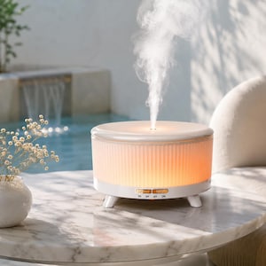 May include: A white cylindrical essential oil diffuser with a ribbed glass exterior that emits a warm orange glow and a steady stream of mist. The base features control buttons labeled LIGHT, MIST, 1H, 3H, 6H, and ON, supported by three small legs.
