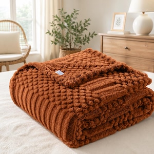 May include: A folded, burnt orange throw blanket featuring a textured, raised popcorn-style knit pattern. The blanket has a small, rectangular white fabric tag attached to the top edge. The material appears soft and plush with a consistent color.