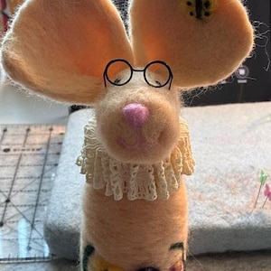 May include: A needle-felted mouse figurine in a light peach color, featuring large ears, a pink nose, and thin black wire glasses. The mouse wears a cream-colored lace ruff collar and has a floral pattern embroidered on its lower body.