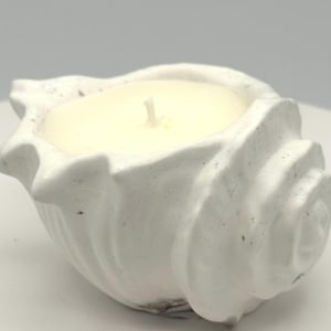 May include: A white, speckled seashell-shaped candle holder containing a cream-colored wax candle with a single centered wick. The exterior of the shell features a textured, matte finish with subtle dark gray flecks throughout the surface.