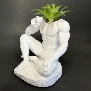 Peut inclure: Un pot de fleurs blanc moucheté en forme de silhouette musclée assise tenant une petite tasse. La figurine repose sur une base texturée et présente une plante succulente verte émergeant du cou, formant un récipient sculptural pour plantes.