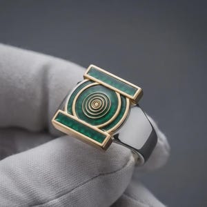以下が含まれることがあります： 銀色のリングで、緑色のエナメルが埋め込まれた長方形の金色のフレームが特徴です。リングの中央には、緑色のエナメルを背景に、同心円状の金色の円形デザインが施されています。