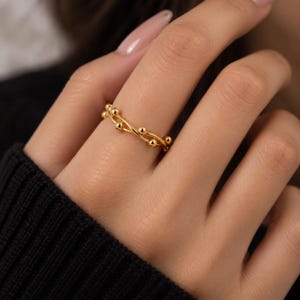 Könnte beinhalten: Ein goldener Ring mit einem zarten, gedrehten Banddesign, das mit kleinen, gleichmäßig verteilten kugelförmigen Perlen entlang des gesamten Umfangs verziert ist. Das Metall hat eine polierte, reflektierende Oberfläche, die das Licht einfängt und ein helles, metallisches Aussehen erzeugt.