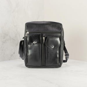May include: A black vertical messenger bag featuring a main zippered compartment, a secondary zippered front pocket, and two smaller front pouches with snap button closures. The bag includes a side strap attachment and silver-toned metal hardware.