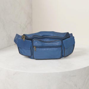 May include: A blue leather fanny pack featuring a large main zippered compartment, a smaller front zippered pocket, and two side pockets with flap closures. The bag includes brass-toned metal zippers and dark blue piping along the edges.