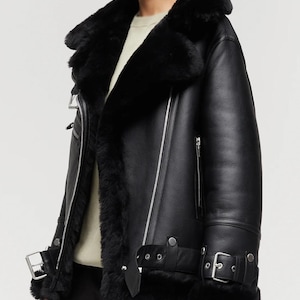 Peut inclure: Une veste en cuir noir avec une doublure et un col en peau lainée noire épaisse. La veste présente une grande fermeture éclair frontale, des détails en métal argenté, des sangles à boucle réglables aux poignets et à la base, et des poches zippées.