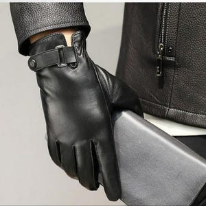 Könnte beinhalten: Ein schwarzer Lederhandschuh mit einem Handgelenkriemen, einer silberfarbenen Metallschnalle und einem Druckknopfverschluss. Der Handschuh weist sichtbare Nähte entlang der Finger und des Handflächenbereichs auf und bietet eine strukturierte Passform.
