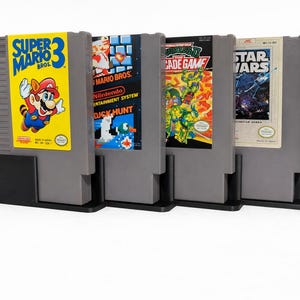 May include: Four gray plastic video game cartridges for the Nintendo Entertainment System, each held upright in a black plastic stand. The labels feature Super Mario Bros. 3, Super Mario Bros./Duck Hunt, Teenage Mutant Ninja Turtles II, and Star Wars.