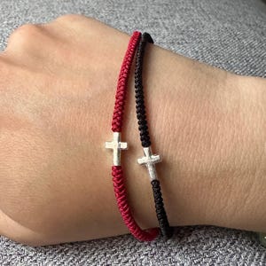May include: Two braided cord bracelets, one in bright red and one in black, each featuring a small, centred silver-toned cross charm. The bracelets are constructed with a woven texture and are positioned side-by-side on a wrist.