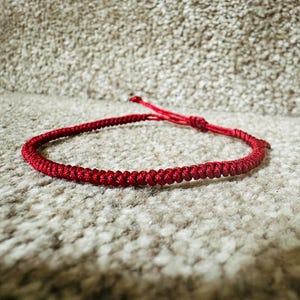 May include: A bright red cord bracelet featuring a continuous braided pattern throughout the band. The bracelet includes an adjustable sliding knot closure with two thin cord ends extending from the centre, creating a simple and functional design.