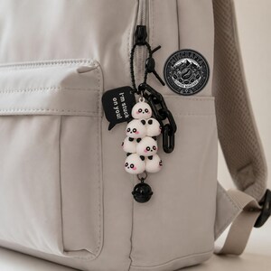 May include: A light gray backpack featuring a hanging keychain accessory. The keychain consists of a stack of five small white panda figures with pink cheeks, a black bell, and a black speech bubble tag with the text I'm stuck on you!