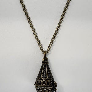 May include: A bronze-toned metal pendant necklace featuring a teardrop-shaped locket with an intricate openwork filigree design. The locket has a hinged side and hangs from a matching bronze-colored link chain with a textured, dark finish.