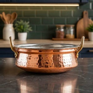 May include: A round, hammered copper serving bowl with a metallic silver-toned interior and two curved brass-coloured handles attached to the sides. The exterior surface features a textured, dimpled finish that reflects light across the metal.