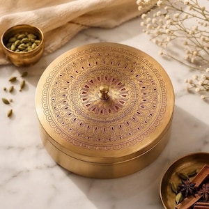Op de afbeelding: Een ronde messing container met een deksel voorzien van een ingewikkeld, gegraveerd mandala-patroon. Het metalen oppervlak heeft een warme, gouden tint met subtiele roodachtige accenten in de geëtste lijnen en een kleine, centrale knop.