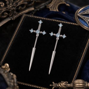 Könnte beinhalten: Ein Paar Ohrringe aus silberfarbenem Metall in Form von Schwertern. Jeder Ohrring verfügt über eine lange, spitze Klinge und ein Parierstange, die mit fünf kleinen, klaren, rund geschliffenen Steinen im Griff und Querbalken besetzt ist.