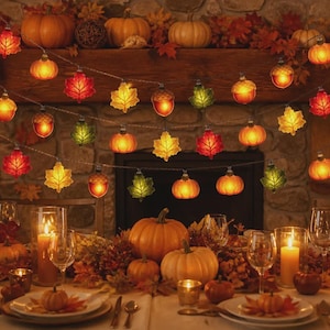 Pode incluir: Um cordão de luzes decorativas com folhas de bordo, abóboras e bolotas translúcidas em tons de laranja, amarelo, vermelho e verde. As luzes estão dispostas em frente a uma lareira de pedra sobre uma mesa com velas e abóboras.