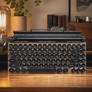 May include: A mechanical keyboard with a wooden base and round, black, typewriter-style keys with white lettering. The keyboard features a black metal top bar with a carriage return lever on the left side and a cylindrical knob on the right side.