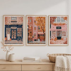 Peut inclure: Trois impressions d'art encadrées représentant des scènes de rue en automne. L'image de gauche montre une librairie avec un vélo, celle du centre des immeubles en briques et un lampadaire, et celle de droite un café avec des citrouilles.