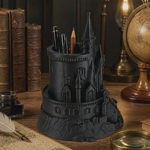 May include: A matte black desk organizer shaped like a castle tower with multiple turrets and architectural details. The cylindrical container holds two pens and a wooden pencil, featuring a textured base that mimics a rocky foundation.