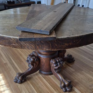 May include: A round wooden table with a dark brown finish and a visible wood grain texture. The base features carved lion head details on each of the four legs. Two rectangular wooden planks with a dark finish are placed on the table surface.