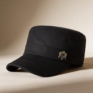 Könnte beinhalten: Eine einfarbige schwarze Militärkappe mit flacher Oberseite und gebogenem Schirm. Die Mütze verfügt über ein horizontales Band um die Basis und ein kleines, metallisches, schildförmiges Emblem an der Seite, das ein detailliertes Wappen zeigt.