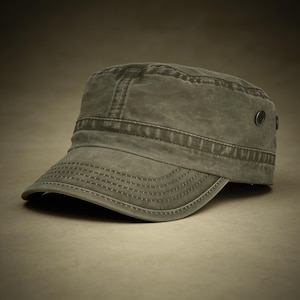 May include: A faded olive green military-style cadet cap featuring a flat top, a short curved brim with multiple rows of decorative stitching, and a small metal grommet on the side panel. The fabric has a worn, textured appearance throughout.