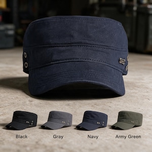 Op de afbeelding: Een marineblauwe militaire cadetpet met een platte bovenkant, een korte gebogen klep en metalen oogjes aan de zijkanten. Een klein rechthoekig metalen logoplaatje is aan de zijkant bevestigd. De afbeelding toont vier kleurvarianten: zwart, grijs, marine en legergroen.