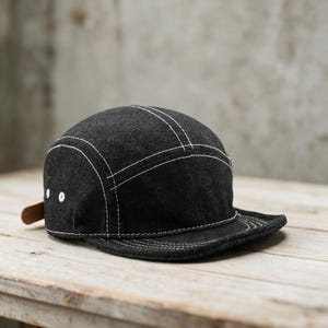 May include: A five-panel cap made of black denim fabric featuring prominent white contrast stitching along the seams and brim. The cap includes two white metal eyelets on the side panel and a small brown leather-like tab at the back closure.