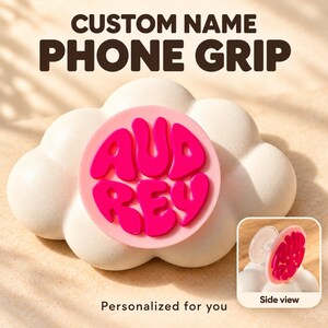 May include: A round phone grip featuring the name AUDREY in raised, bright pink, wavy-style lettering on a light pink circular base. A smaller inset shows a side view of the clear plastic collapsible mount attached to the back.