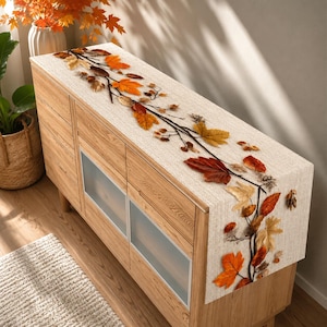 May include: A rectangular table runner made of light beige fabric featuring a printed design of dark brown branches adorned with vibrant autumn leaves in shades of orange, red, yellow, and brown, arranged in a flowing pattern across the surface.