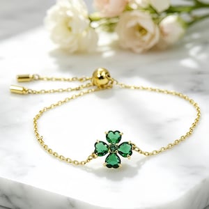 May include: A gold-tone chain bracelet featuring a central four-leaf clover charm composed of four heart-shaped green gemstones and a small central green stone. The bracelet includes an adjustable sliding bead mechanism with two gold-tone end caps.