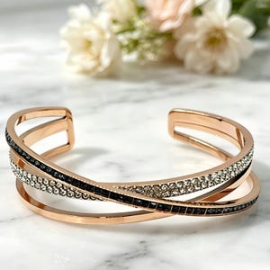 Könnte beinhalten: Ein roségoldfarbenes Armband in Manschettenform mit einem mehrschichtigen, überkreuzten Design. Das Armband ist mit zwei Reihen eingebetteter Kristalle verziert, wobei eine Reihe klare weiße Steine und die andere kleine schwarze Steine enthält.