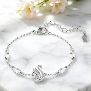 May include: A silver-toned chain bracelet featuring a central swan-shaped charm encrusted with clear crystals. The bracelet includes four additional marquise-cut crystal accents along the chain, a lobster claw clasp, and a small octagonal logo tag.