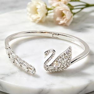 May include: A silver-toned hinged bangle bracelet featuring a swan-shaped motif encrusted with clear, sparkling stones. One side of the open-ended design displays a row of five oval-cut stones, while the other side showcases a swan silhouette.