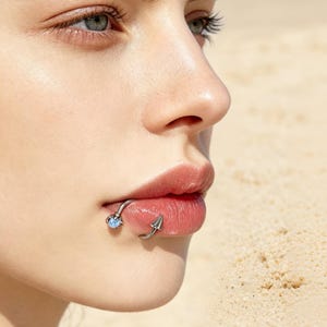 May include: A silver-toned horseshoe-shaped lip ring featuring a small, round, iridescent blue stone held by a claw setting on one end and a pointed, cone-shaped stud on the other end, positioned on the lower lip of a face.