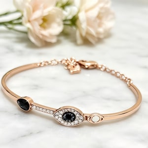 Könnte beinhalten: Ein roségoldfarbenes Armband mit einem zentralen, augenförmigen Anhänger, der mit klaren Kristallen und einem schwarzen Stein besetzt ist, einem tropfenförmigen schwarzen Stein, einem kleinen offenen Kreisdetail und einer feinen Kette mit Karabinerverschluss.