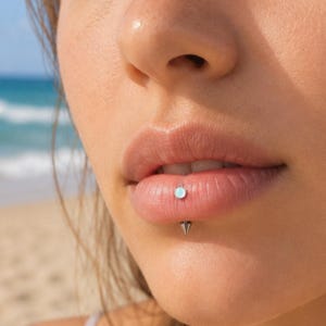 Puede incluir: Una joya de piercing labial en tono plateado que presenta una pequeña piedra circular iridiscente en la superficie exterior y un perno puntiagudo en forma de cono que sobresale del borde inferior del labio, colocado verticalmente.