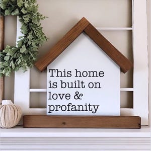 May include: White wooden house-shaped sign with the text "This home is built on love & profanity". The sign is sitting on a wooden shelf.