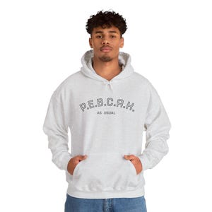 Peut inclure: Un sweat-shirt à capuche gris chiné clair avec une poche kangourou sur le devant et des manches longues. La poitrine présente le texte noir P.E.B.C.A.K. en police arquée, avec les mots AS USUAL imprimés en plus petit juste en dessous.