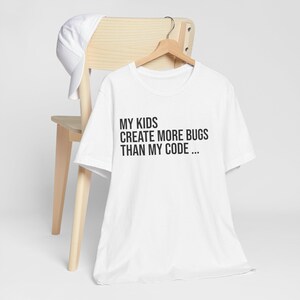 Op de afbeelding: Een wit T-shirt met korte mouwen en de zwarte tekst "MY KIDS CREATE MORE BUGS THAN MY CODE ..." op de voorzijde. Het T-shirt hangt aan een houten kledinghanger en een effen witte baseballpet ligt over de rugleuning van de stoel.