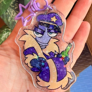 May include: A clear acrylic keyring featuring a cartoon character wearing a purple police hat with a yellow star and a purple coat with tan fur trim. The character holds a green bottle and the keyring has a purple star-shaped plastic clasp.