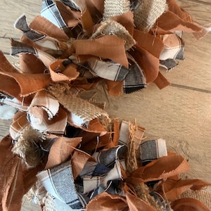 May include: A decorative garland constructed from numerous tied fabric strips in shades of burnt orange, tan, and white, interspersed with black and white plaid patterns and textured natural burlap pieces with frayed edges.