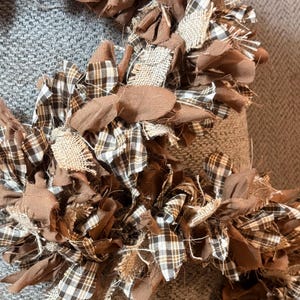 May include: A circular rag wreath constructed from numerous strips of fabric tied together. The fabric pieces feature a mix of solid brown cotton, brown and white plaid patterns, and textured tan burlap, creating a layered, frayed appearance.