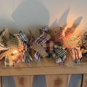 May include: A decorative garland featuring a mix of fabric strips in red, green, and blue plaid patterns interspersed with natural tan burlap strips. The garland is illuminated by small, warm-toned string lights woven throughout the fabric pieces.
