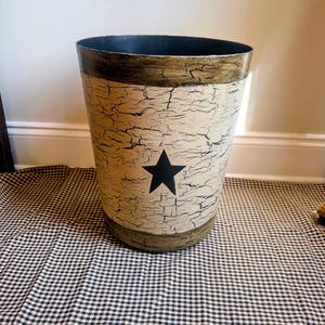 May include: A cylindrical wastebasket featuring a cream-colored center with a black crackle finish and a solid black star emblem in the middle. The top and bottom rims are finished in a dark brown, textured crackle paint, creating a contrast.