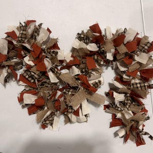 May include: A candy cane shaped rag wreath constructed from tied fabric strips in a color palette of burnt orange, cream, and brown plaid, interspersed with textured tan burlap strips, creating a full and frayed appearance throughout the design.