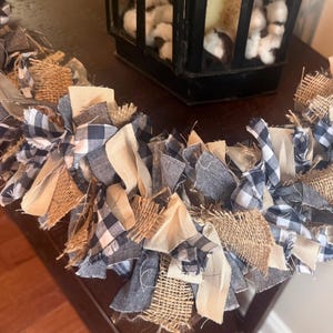 May include: A decorative rag garland constructed from strips of fabric tied together. The garland features a mix of textures and patterns, including tan burlap, solid cream fabric, solid dark blue fabric, and a dark blue and white gingham check.