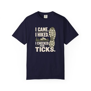 May include: A navy blue short-sleeve t-shirt featuring a graphic print with the text "I CAME. I HIKED. I CHECKED FOR TICKS." alongside an illustration of a mountain range, pine trees, and a large boot print with a small tick icon inside the heel.