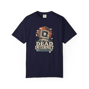 May include: A navy blue short-sleeve t-shirt featuring a graphic of a vintage computer monitor with a skull on the screen, a crow perched on top, and the text "DEAD INTERNET SOCIAL CLUB" printed in a beige font below the computer illustration.